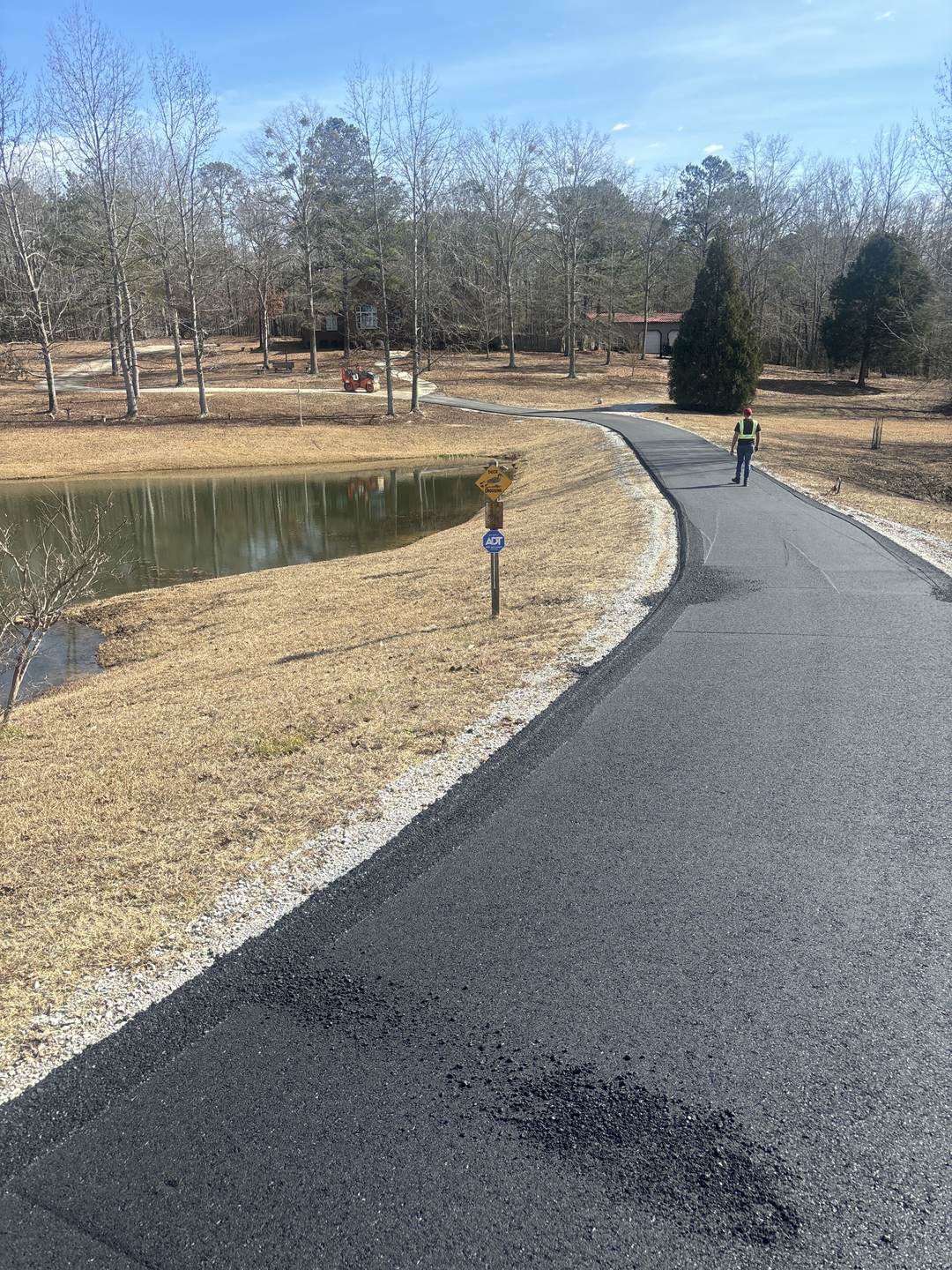 The same pond lane after paving, fresh black asphalt curving past the water
